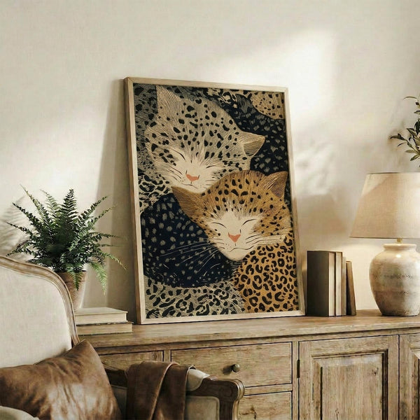 May include: Framed poster displaying two stylized cats with spotted leopard-like patterns in golden yellow and dark blue tones, positioned on rustic wooden sideboard alongside fern plant, ceramic vase, and books in neutral-toned living room setting