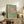 May include: Framed art print with sage green textured background featuring pink circle sun and silhouetted birds in flight, displayed on rustic wood console table alongside fern plant, ceramic vases, and stacked books in neutral-toned living room setting with cream upholstered furniture.