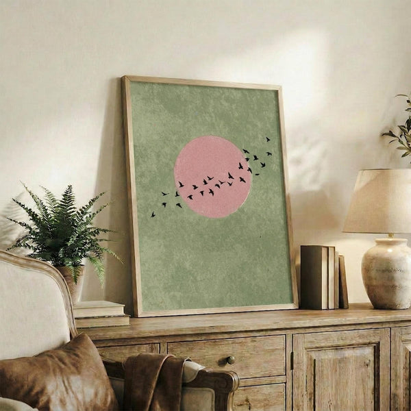May include: Framed art print with sage green textured background featuring pink circle sun and silhouetted birds in flight, displayed on rustic wood console table alongside fern plant, ceramic vases, and stacked books in neutral-toned living room setting with cream upholstered furniture.