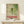May include: Vintage-style poster featuring large green dragonfly with transparent wings against soft green cloudy background, Japanese text on left side, red "TOKYO" lettering at bottom, "JAPAN 1977" and "DRAGONFLY" text, displayed in thin wooden frame on light wood floating shelf with small white wrapped gift box beside it, against cream colored wall