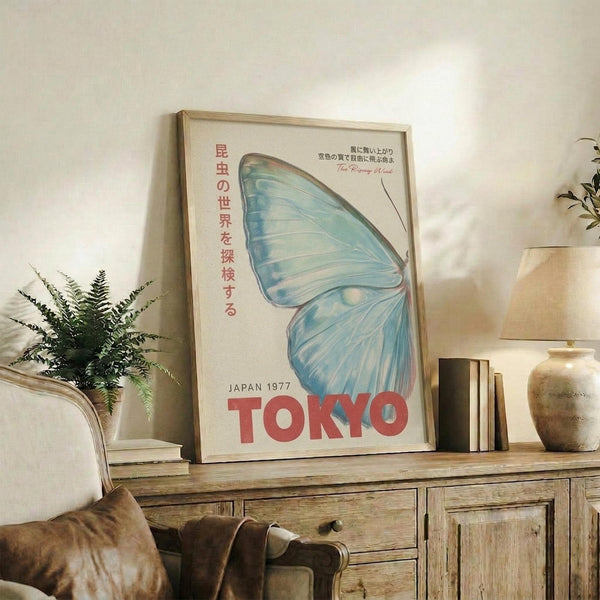May include: Framed vintage-style poster featuring blue butterfly wing design with Japanese text and red "TOKYO" lettering, displayed on rustic wooden sideboard alongside fern plant, ceramic vases, and books in cozy living room setting with cream armchair and neutral walls.