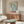 May include: Vintage-style poster featuring large blue butterfly wing design with Japanese text and red "TOKYO" lettering, displayed in thin frame above mid-century walnut credenza with brass hardware. Modern white sculptural chair, glass-top oval coffee table, brass floor lamp, and small white table lamp complete the minimalist room setting with neutral walls and natural lighting.