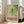 May include: Framed vintage-style poster with large green dragonfly illustration on weathered green background, featuring Japanese text in red characters and "TOKYO" in bold red letters, dated "JAPAN 1977" and "1977年4月15日". Light wood frame leans against white wall next to beige upholstered chair in bright, naturally-lit room with white window frames.