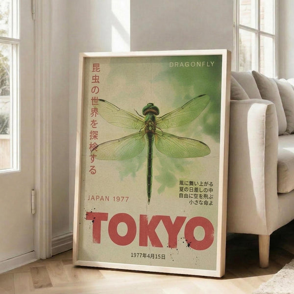 May include: Framed vintage-style poster with large green dragonfly illustration on weathered green background, featuring Japanese text in red characters and "TOKYO" in bold red letters, dated "JAPAN 1977" and "1977年4月15日". Light wood frame leans against white wall next to beige upholstered chair in bright, naturally-lit room with white window frames.