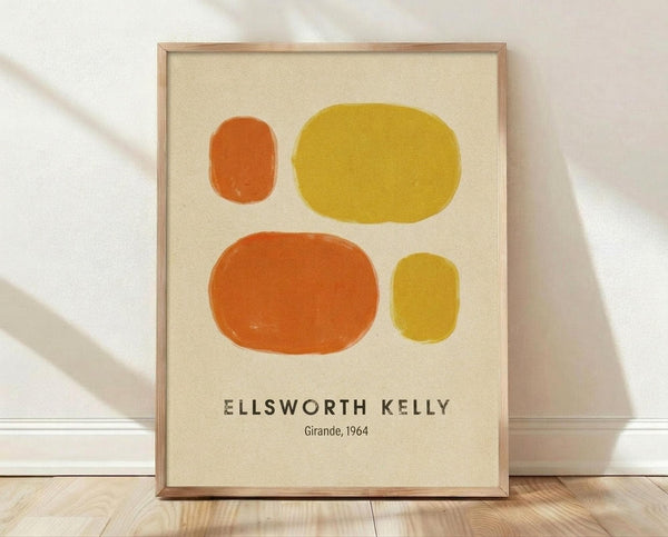 May include: Framed art print leaning against white wall on light wood floor. Cream-colored poster features four abstract oval shapes in orange and yellow tones - small orange oval top left, large yellow oval top right, large orange oval bottom left, small yellow oval bottom right. Black text at bottom reads "ELLSWORTH KELLY" and "Girande, 1964". Natural wood frame with soft shadows from window light creating diagonal patterns across the scene.