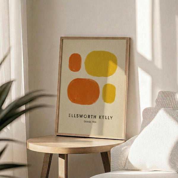 May include: Framed abstract art print featuring four organic oval shapes in orange and yellow tones on cream background, displayed on round wooden side table next to beige upholstered chair with white pillow. Natural sunlight creates soft shadows across white walls, with green plant leaves visible in corner of minimalist room setting.
