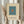 May include: Two hands holding a framed minimalist art print featuring a small orange square centered on a teal blue background with cream matting and light wood frame. Person wearing beige clothing with puffy sleeves holds dried pampas grass in one hand against a neutral backdrop with soft natural lighting.