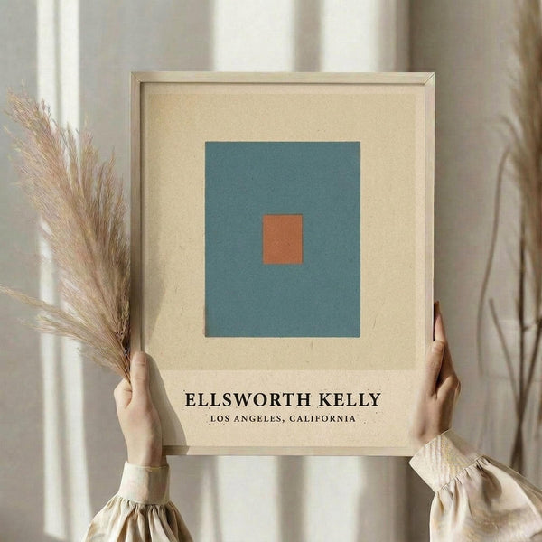 May include: Two hands holding a framed minimalist art print featuring a small orange square centered on a teal blue background with cream matting and light wood frame. Person wearing beige clothing with puffy sleeves holds dried pampas grass in one hand against a neutral backdrop with soft natural lighting.