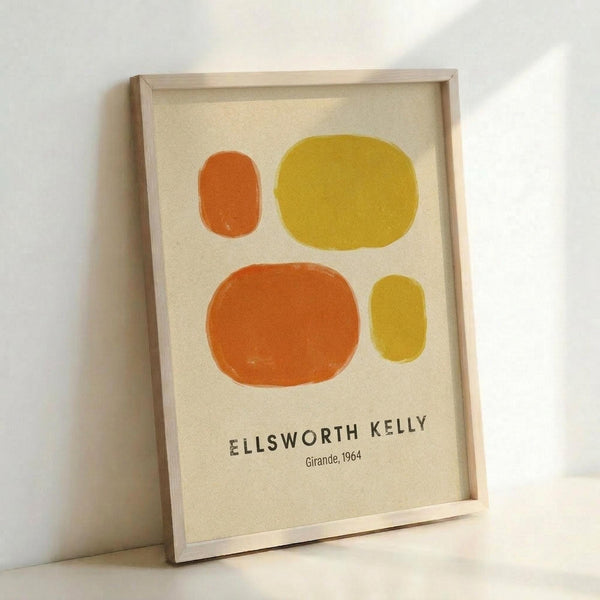 Framed art print featuring four abstract oval shapes in orange and yellow tones on a cream background. The minimalist geometric composition shows two larger ovals and two smaller ones arranged asymmetrically. Text below reads "ELLSWORTH KELLY" and "Girande, 1964" in dark letters. The print is displayed in a light wood frame leaning against a white wall.