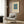 May include: Beige armchair with geometric patterned pillow positioned next to framed art print on wood-paneled wall. Print features teal blue square with small orange square center on cream background with black text below. Gold floor lamp and dark round side table with open book complete the modern living room mockup scene.