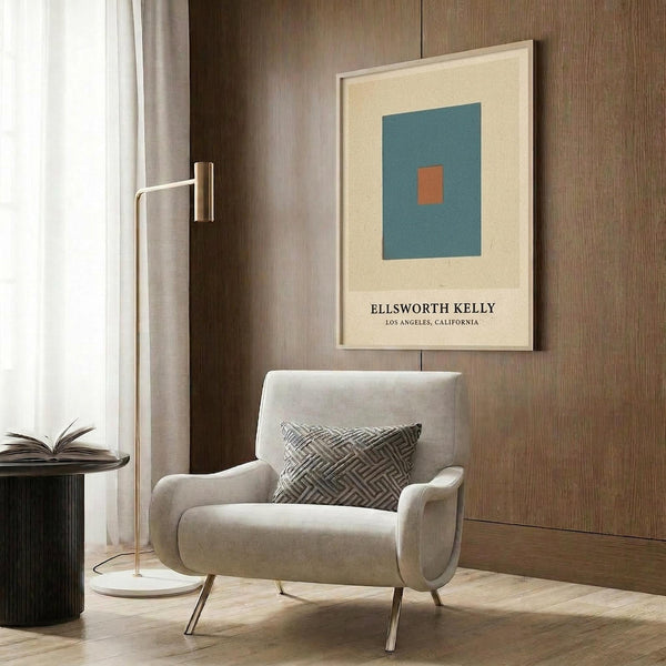 May include: Beige armchair with geometric patterned pillow positioned next to framed art print on wood-paneled wall. Print features teal blue square with small orange square center on cream background with black text below. Gold floor lamp and dark round side table with open book complete the modern living room mockup scene.