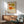 May include: Framed art print featuring four abstract oval shapes in orange, yellow, and olive green on cream background, displayed above mid-century modern wooden credenza with table lamp. Mint green upholstered chair with wooden arms sits nearby on hardwood floor. Sunlight creates geometric shadows on white wall in minimalist living room setting.