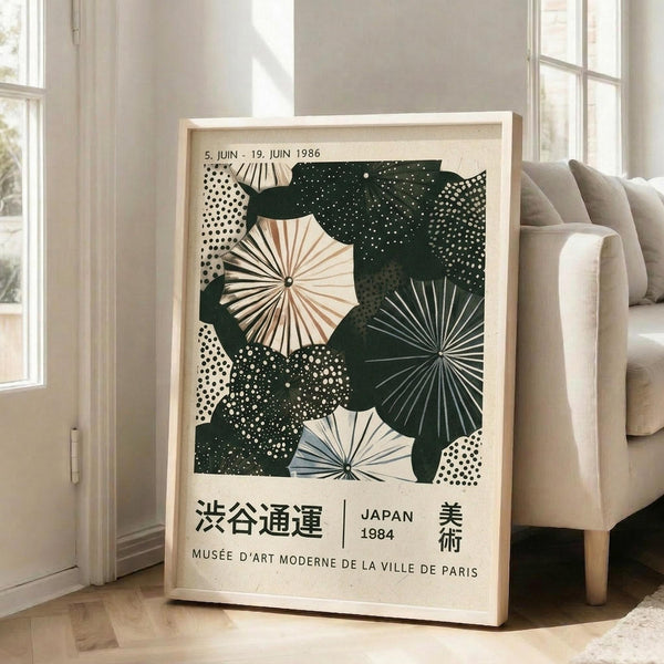 May include: A framed vintage exhibition poster featuring abstract umbrellas and circular patterns in forest green, cream, and light blue tones, displayed on a light wooden floor against white walls near a beige upholstered chair and tall windows with natural lighting.