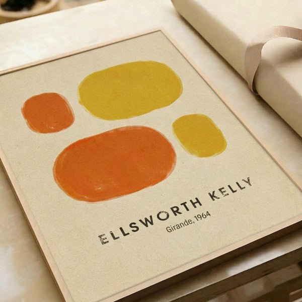 May include: Framed art print featuring four abstract oval shapes in orange and yellow tones on cream background, displayed on white surface next to rolled paper tube. Print shows "ELLSWORTH KELLY" and "Girande, 1964" text at bottom in black letters within thin border frame.