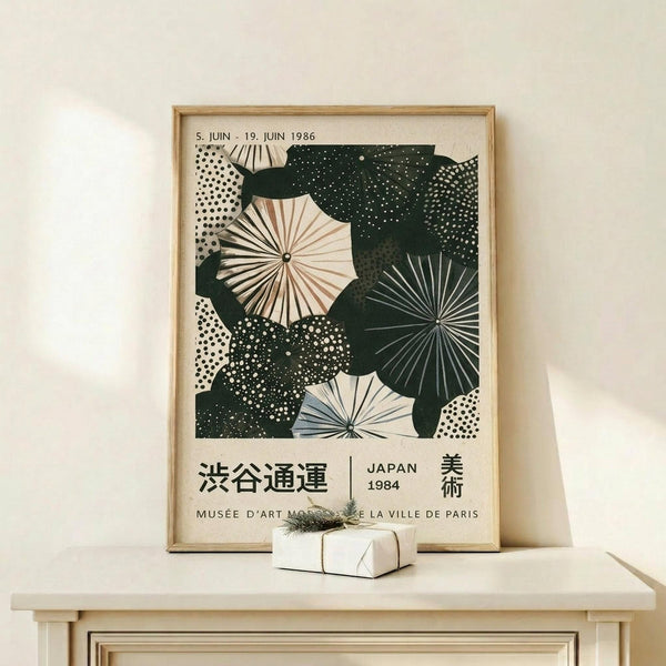 May include: Framed vintage-style poster featuring green and cream abstract umbrella shapes with dotted patterns and radiating lines, displayed on light wood shelf next to small white wrapped gift with evergreen sprig. Japanese and French text visible at bottom. Natural lighting creates soft shadows on cream wall background.