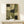 May include: Abstract geometric artwork in earth tones displayed in thin wooden frame on light wood surface against cream wall. Composition features overlapping curved and angular shapes in olive green, sage, cream, and dark forest tones with textured, weathered appearance. Small wrapped white gift box with natural twig and feather sits beside the framed print.