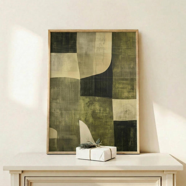 May include: Abstract geometric artwork in earth tones displayed in thin wooden frame on light wood surface against cream wall. Composition features overlapping curved and angular shapes in olive green, sage, cream, and dark forest tones with textured, weathered appearance. Small wrapped white gift box with natural twig and feather sits beside the framed print.