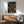 May include: Framed Japanese street scene artwork displayed above wooden sideboard in modern living room. Print shows narrow alley with stone steps, glowing red and orange paper lanterns, traditional buildings with Japanese signage, rendered in deep blues and warm lighting. Mid-century furniture includes teal upholstered armchair and white table lamp.