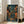 May include: Framed artwork leaning against white wall next to beige sofa in bright room with large windows. The poster shows a moody Japanese street scene at night with glowing red lanterns, wooden stairs leading upward, traditional shop signs with Japanese text, and wet cobblestone ground reflecting warm amber and orange lights. The artwork has rich teal and blue tones contrasted with warm lighting, displayed in a light wood frame.
