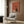 May include: Framed retro-style poster featuring black cat silhouette with white eyes sitting beside white coffee pot and cup on orange-red background, displayed on wood-paneled wall above beige upholstered armchair with geometric patterned pillow, alongside brass floor lamp and dark side table with open book in modern living room setting with neutral tones and natural lighting.