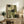 May include: Abstract geometric artwork in earth tones with green, cream, and beige rectangular shapes displayed in light wood frame on rustic wooden sideboard alongside fern plant, ceramic vase, and books in warm, naturally-lit living room setting.