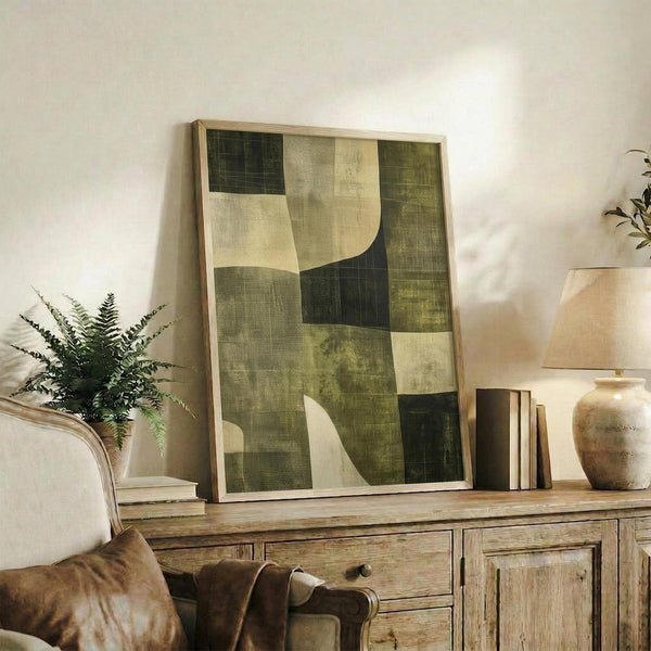 May include: Abstract geometric artwork in earth tones with green, cream, and beige rectangular shapes displayed in light wood frame on rustic wooden sideboard alongside fern plant, ceramic vase, and books in warm, naturally-lit living room setting.