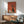 May include: Framed vintage-style poster with orange-red background featuring minimalist black cat silhouette sitting beside white coffee pot and cup, displayed above mid-century modern wooden sideboard with table lamp, next to sage green upholstered armchair with wood arms in sunlit room with white walls.