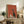 May include: Framed retro-style poster featuring a tall black cat with wide white eyes sitting on a surface next to white coffee pot and cup against terracotta orange background. Light wood frame displayed on rustic wooden sideboard with books, ceramic vase, and fern plant in cozy living room setting with beige armchair.
