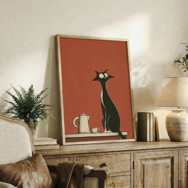 May include: Framed retro-style poster featuring a tall black cat with wide white eyes sitting on a surface next to white coffee pot and cup against terracotta orange background. Light wood frame displayed on rustic wooden sideboard with books, ceramic vase, and fern plant in cozy living room setting with beige armchair.