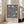 May include: Framed poster with navy blue background featuring ornate floral pattern in cream and coral colors, displaying tulip-like blooms and decorative leaves in William Morris style. Text at bottom reads "Centenary Exhibition William Morris" with exhibition details. Poster leans against white wall beside beige upholstered chair in bright, naturally-lit room with hardwood floors.