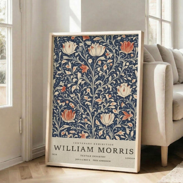May include: Framed poster with navy blue background featuring ornate floral pattern in cream and coral colors, displaying tulip-like blooms and decorative leaves in William Morris style. Text at bottom reads "Centenary Exhibition William Morris" with exhibition details. Poster leans against white wall beside beige upholstered chair in bright, naturally-lit room with hardwood floors.