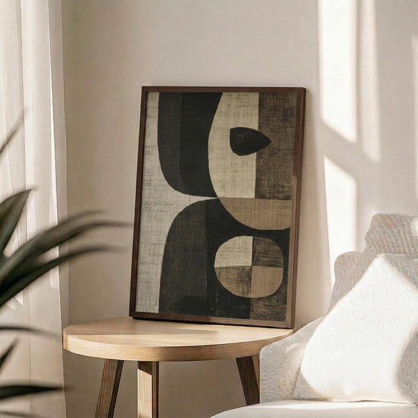 May include: Framed abstract art print featuring geometric shapes in dark green and cream tones with textured, weathered appearance. Print sits on round wooden side table next to beige upholstered chair with white pillow. Soft natural lighting creates shadows on neutral wall, with plant leaves visible in corner of modern living space.