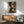 May include: Modern living room with sage green upholstered armchair, walnut sideboard, and white table lamp. Large framed abstract art print on white wall features geometric shapes in dark green, cream, and brown with textured, weathered appearance. Natural sunlight streams across the space, highlighting the mid-century modern furniture and warm wood tones throughout the room.