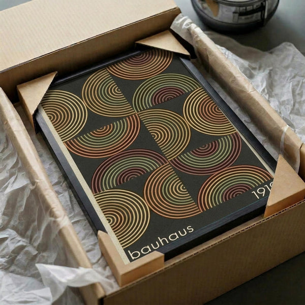 May include: Framed geometric art print featuring overlapping semicircular shapes in warm autumn colors - rust red, golden yellow, and olive green - on a dark background. The print sits in a wooden frame inside cardboard packaging with tissue paper, surrounded by wooden dowels and shipping materials on a desk surface.