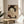 May include: Framed Japanese-style artwork displaying a geisha with black hair adorned with white flowers, holding an ornate fan that partially covers her face, set against a decorative wave pattern background. The print sits on a light wooden round side table next to a beige upholstered chair with white pillows, near a window with natural lighting and a potted plant visible in the corner.