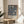 May include: Framed vintage-style exhibition poster featuring William Morris floral textile pattern in navy blue with cream and coral tulips and leaves, displayed on light wood round side table next to beige upholstered chair, with plant leaves visible and natural window lighting creating soft shadows on neutral wall.