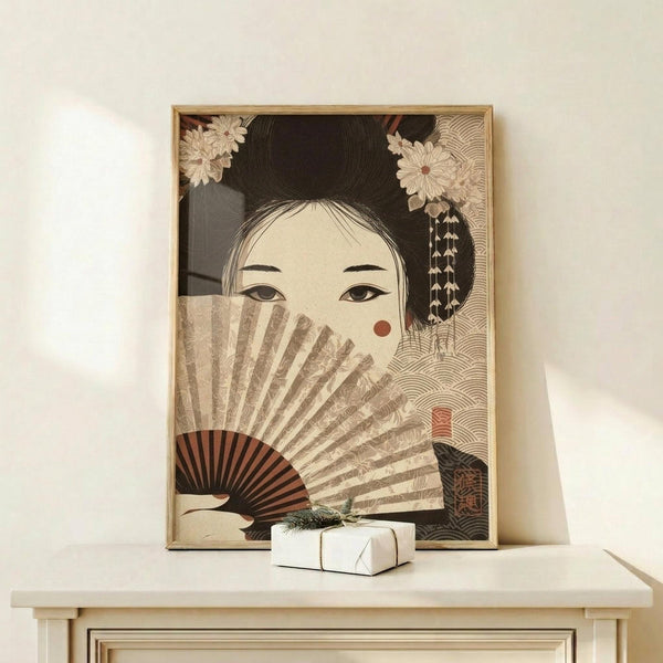 May include: Framed vintage-style Japanese art print featuring a geisha with elaborate black hair adorned with white chrysanthemum flowers and dangling ornaments. She holds a decorative fan partially covering her face, with almond-shaped eyes and a small red beauty mark visible. The artwork uses muted earth tones with intricate wave patterns in the background. Displayed on a light wood console table with a small white wrapped gift, against a cream colored wall.
