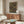 May include: Modern living room with white curved chair and oval glass coffee table. Wood credenza displays framed geometric poster featuring overlapping circles in earth tones - rust, olive, cream on dark background with 1919 Bauhaus text. Contemporary table lamp and floor lamp complete the mid-century modern styling against neutral walls.