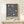 May include: Framed William Morris exhibition poster featuring blue background with ornate floral pattern in cream, coral and orange tulips and flowing botanical motifs. Wood frame sits on light wooden shelf beside small wrapped white gift box with evergreen sprig against neutral wall.