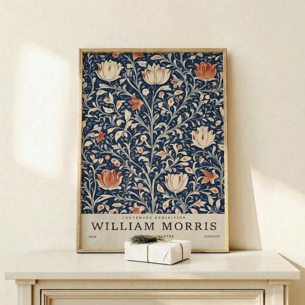 May include: Framed William Morris exhibition poster featuring blue background with ornate floral pattern in cream, coral and orange tulips and flowing botanical motifs. Wood frame sits on light wooden shelf beside small wrapped white gift box with evergreen sprig against neutral wall.