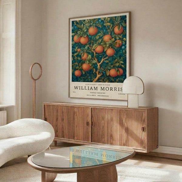 May include: William Morris exhibition poster featuring orange fruit tree pattern on blue-green background, displayed in modern living room with walnut credenza, white sculptural chair, glass coffee table, and brass floor lamp. Poster shows vintage botanical design with oranges and foliage in ornate Arts and Crafts style, framed in light wood against cream wall.