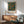 May include: Framed vintage-style exhibition poster featuring orange fruit tree with lush green foliage against deep blue background, displayed above mid-century modern wooden credenza with white ceramic lamp, next to sage green upholstered armchair with wooden frame in sunlit room with white walls.