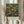 May include: Hands holding a framed vintage exhibition poster featuring an orange tree with abundant fruit against a deep teal background in William Morris's distinctive Arts and Crafts style. The botanical illustration shows detailed leaves and branches with ripe oranges throughout. Below the artwork is cream-colored text reading "CENTENARY EXHIBITION WILLIAM MORRIS TEXTILE INDUSTRY" with exhibition dates. Dried pampas grass sits nearby against soft cream curtains in natural lighting.