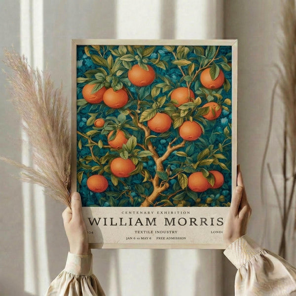 May include: Hands holding a framed vintage exhibition poster featuring an orange tree with abundant fruit against a deep teal background in William Morris's distinctive Arts and Crafts style. The botanical illustration shows detailed leaves and branches with ripe oranges throughout. Below the artwork is cream-colored text reading "CENTENARY EXHIBITION WILLIAM MORRIS TEXTILE INDUSTRY" with exhibition dates. Dried pampas grass sits nearby against soft cream curtains in natural lighting.