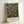 May include: Framed vintage exhibition poster featuring William Morris botanical design with orange fruit tree against deep teal blue background. White text at bottom reads "Centenary Exhibition William Morris London Textile Industry 1934 Jan 6 to May 6 Free Admission." Light wood frame leaning against white wall with soft natural lighting creating gentle shadows.