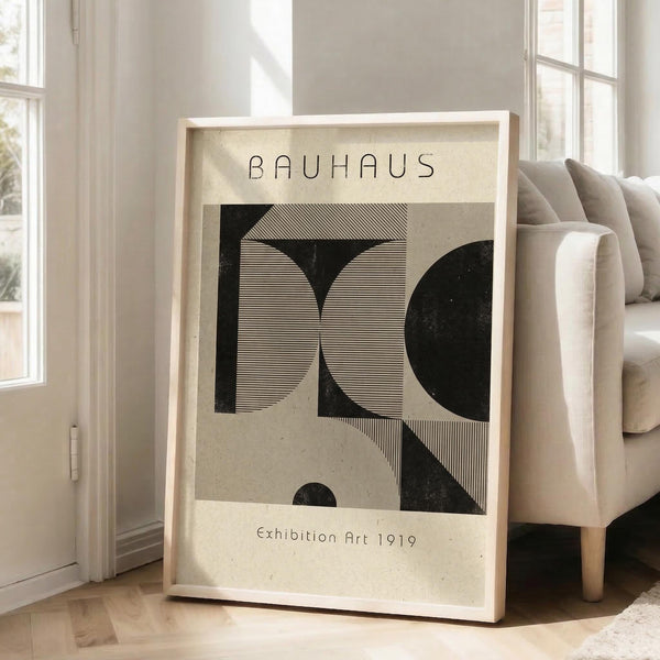 May include: A framed Bauhaus-style poster with geometric black and beige shapes and fine line patterns, displaying "BAUHAUS" and "Exhibition Art 1919" text. The cream-colored artwork sits against a light wood frame on hardwood floors beside a beige upholstered chair near bright windows in a minimalist white room.