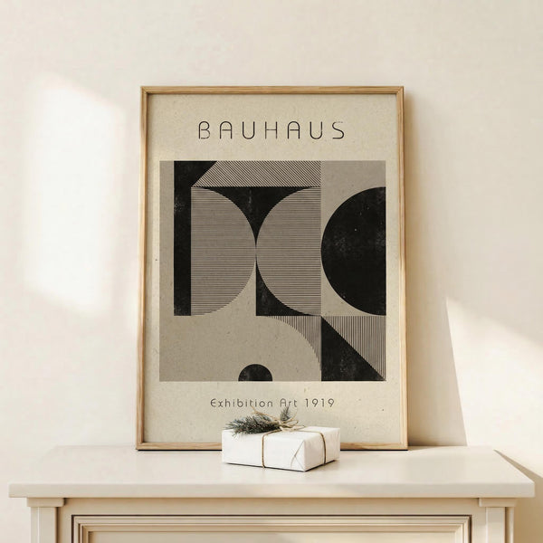 May include: Framed Bauhaus-style poster with geometric black and white abstract design featuring curved and angular shapes with fine line details, displayed on light wood surface alongside small wrapped gift with twine and dried botanicals against neutral wall