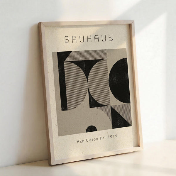 May include: Framed Bauhaus-style art print leaning against white wall in light wood frame. Black geometric shapes with fine line patterns on cream background. Abstract angular forms create dynamic composition. Modern minimalist design with "Exhibition Art 1919" text below artwork. Natural lighting creates soft shadows on textured paper surface.