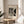 May include: Framed Bauhaus-style abstract art print in thin wood frame, displayed on round wooden side table next to beige upholstered chair. Print features geometric black and white shapes with hatched patterns on cream background. Soft natural lighting creates shadows on neutral wall, with plant leaves visible in corner of minimalist modern room setting.