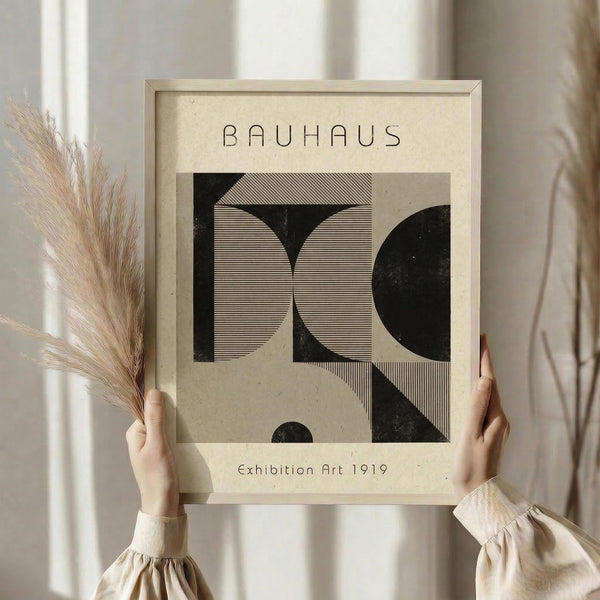 May include: Hands holding a framed Bauhaus-style art print featuring geometric black shapes with fine line patterns on cream background. Person wearing beige clothing holds dried pampas grass, standing against soft curtained window with natural lighting creating warm, minimalist aesthetic.